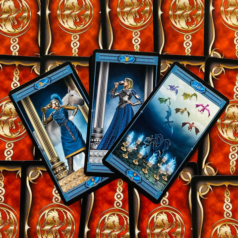Tarot kortos su mėlynais rėmeliais, trys atverstos kortos (karžygė, dama, dešimt taurų) ant raudono auksuoto nugarėlių fono