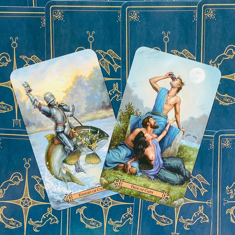 Tarot kortos, dvi atverstos kortos (Riteris taurėje ir Puslapis taurėje) ant mėlyno ornamentuoto fono, parodytos iš viršaus