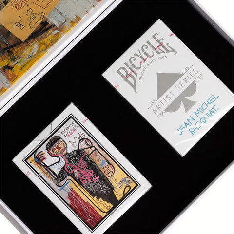 Bicycle kortos Artist Series Jean‑Michel Basquiat baltame pakelyje su iliustruotomis nugarėlėmis juodame pristatymo dėkle, pasvirusios kampu