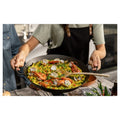 Paella keptuvė 38 cm juoda, patiekiama su krevetėmis ir midijomis, laikoma dviem rankomis ant stalo