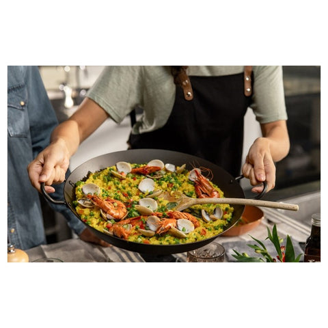 Paella keptuvė 38 cm juoda, patiekiama su krevetėmis ir midijomis, laikoma dviem rankomis ant stalo