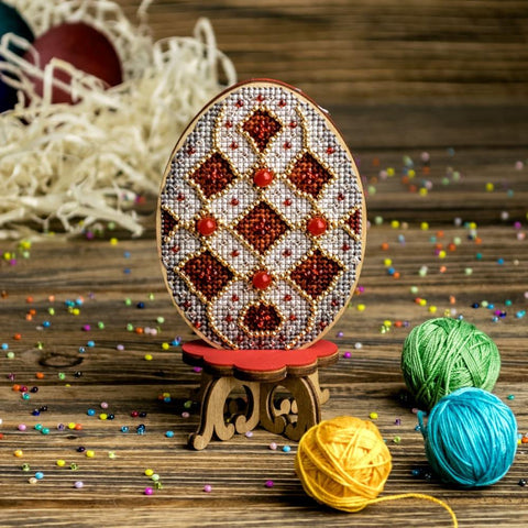 Rankų darbo siuvinėtas kiaušinis su raudonai‑auksine karoliukų ornamentika ant medinio stovelio, nufotografuotas iš priekio, šalia verpalų ir karoliukų.