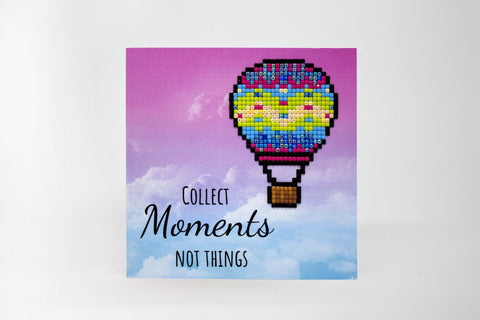 Plakatas su ryškiai rausvai‑mėlynu fonu ir pikseliniu karšto oro baliono motyvu bei įkvepiančiu užrašu Collect Moments Not Things