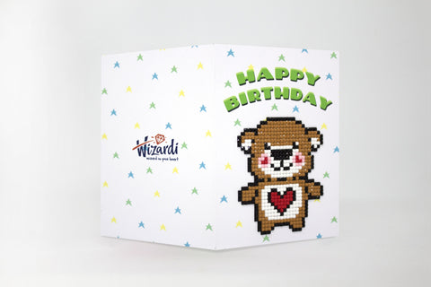 Gimtadienio atvirukas su Wizardi logotipu, baltas žvaigždėtas, stovi atverstais kraštais, pikselinis meškutis su raudona širdimi ir žalia užraša "Happy Birthday" viršuje