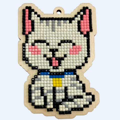 Katės pakabukas iš mažų permatomų karoliukų ant medinės plokštelės, balta „pikselinė“ figūrėlė su mėlyna apykakle ir geltonu pakabuku, priekio artimas planas