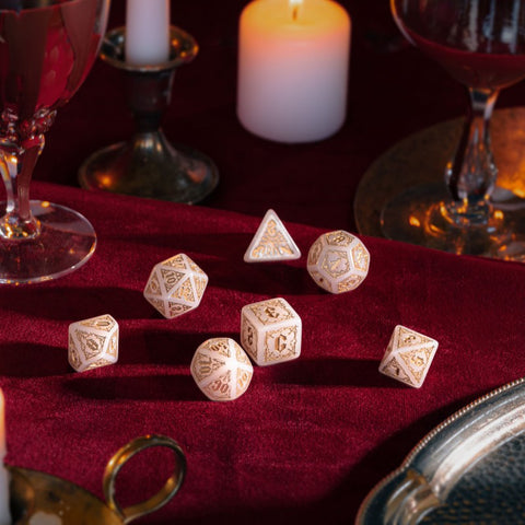 RPG kauliukai baltos spalvos su auksiniais ornamentais, 7 vnt., pateikti ant bordo aksomo šilto žvakių apšvietimo fone