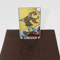 Tarot korta The Fool, Rider–Waite stiliaus, stačia ant mažo medinio stovo, eksponuota ant rudo Fantasy Puzzles kilimėlio, matomas priekis