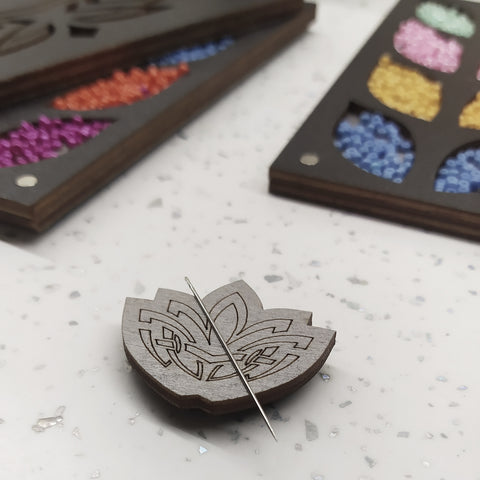 Magnetinis adatos laikiklis lotus formos, tamsaus medžio graviruotė, adata prisegta priekyje, fone spalvoti karoliukų skyreliai