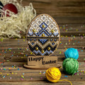 Rankų darbo medinis stovelis su karoliukais dekoruotu Velykų kiaušiniu (mėlyna–auksinė–balta), priekyje „Happy Easter“ plokštelė ir spalvoti siūlų kamuoliukai, artimas planas