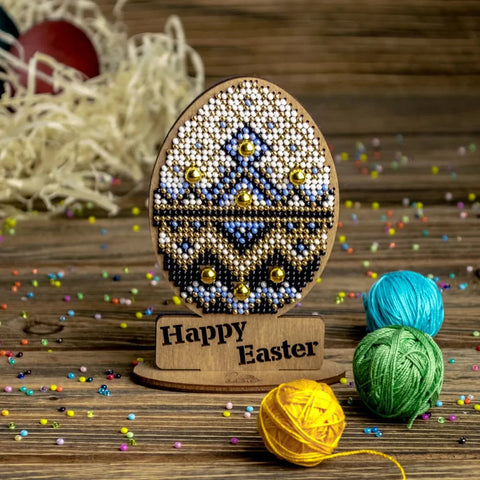 Rankų darbo medinis stovelis su karoliukais dekoruotu Velykų kiaušiniu (mėlyna–auksinė–balta), priekyje „Happy Easter“ plokštelė ir spalvoti siūlų kamuoliukai, artimas planas