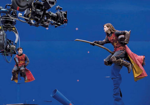 Gryffindor kostiumas raudonas su auksiniais akcentais, ant šluotos skriejantis aktorės kostiumas filmavimo aikštelėje su mėlynu fonu