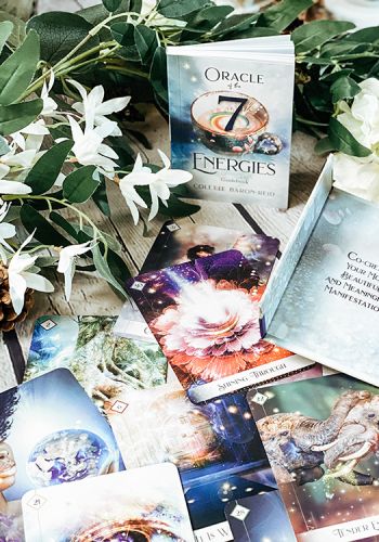 orakulo kortų rinkinys su dėklu "Oracle of the 7 Energies" ir išskleistu kortų komplektu, padėtas ant žalių lapų ir baltų žiedų