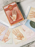 palmistry kortos: oranžinė 64 kortų dėžutė su išskleista pastelinėmis kortomis, rodančiomis delnų linijas