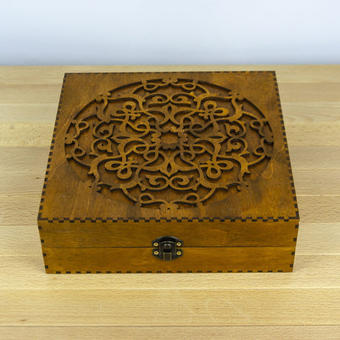 Medinė dėžutė rudos spalvos, apie 20×20 cm, su lazeriu išpjauta ornamentu ant dangčio ir metaliniu užsegimu, matoma iš viršaus ir priekio