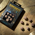 RuneQuest kauliukų rinkinys 7 vnt., kreminės spalvos su bordo simboliais, pateiktas dėžutėje ir išmėtytas ant smėlio