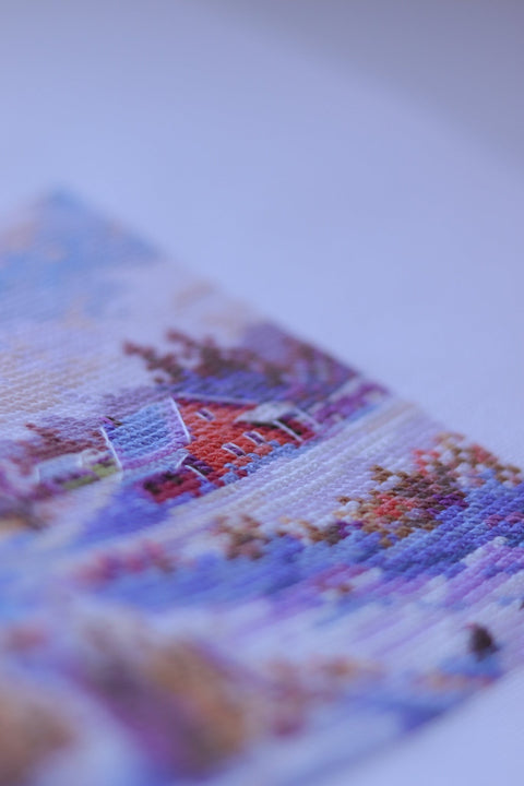 Kryželių siuvinys per arti, kampu rodoma smulkiai matoma tekstūra violetinių ir mėlynų atspalvių su raudonu nameliu