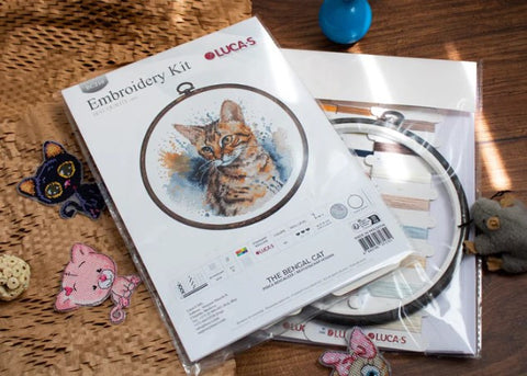 Siuvinėjimo rinkinys Lucas The Bengal Cat su bengalų katės piešiniu, uždaru plastiko pakuotėje, matomu apvaliu lankeliu ir spalvotais siūlais, šalia siuvinėtos dekoracijos.