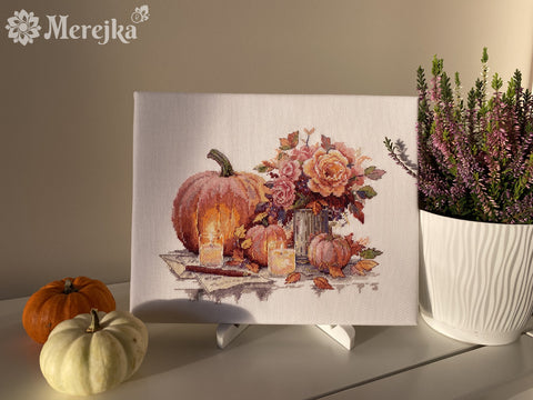 Siuvinėtas paveikslas Merejka su moliūgais, žvakėmis ir rudens gėlių puokšte, stilingai pastatytas ant stalo tarp mažų moliūgų