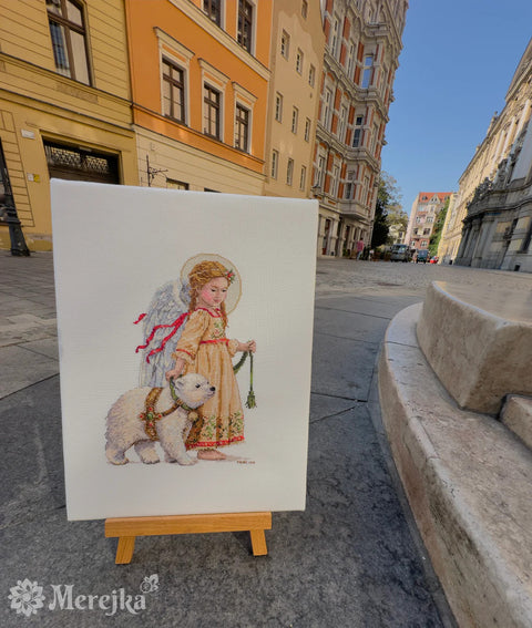Siuvinėtas paveikslas su angelu ir baltu lokiu ant drobės, smulkus kryželių darbas, eksponuotas ant medinio stovelio miesto gatvėje