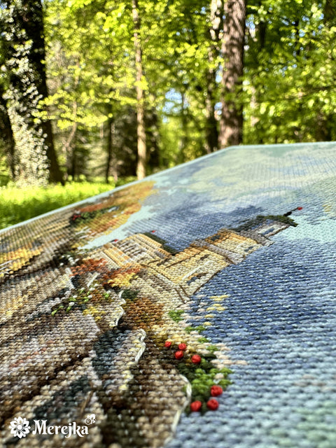 Siuvinys kryželiu Merejka, arti nufotografuotas pasviru kampu su ryškia tekstūra ir raudonomis mazgelinėmis detalėmis, miško fone