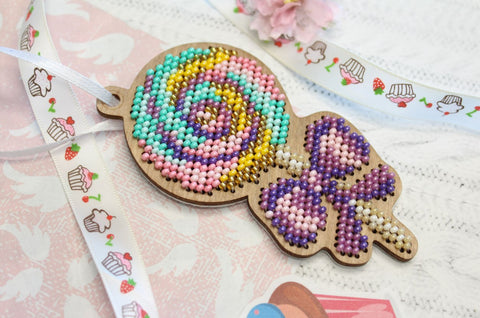 Medinis pakabukas saldainis su pastelinėmis (turkio, rožinė, geltona, violetinė) karoliukų spiralėmis ir rožiniu kaspinu, artimas planas