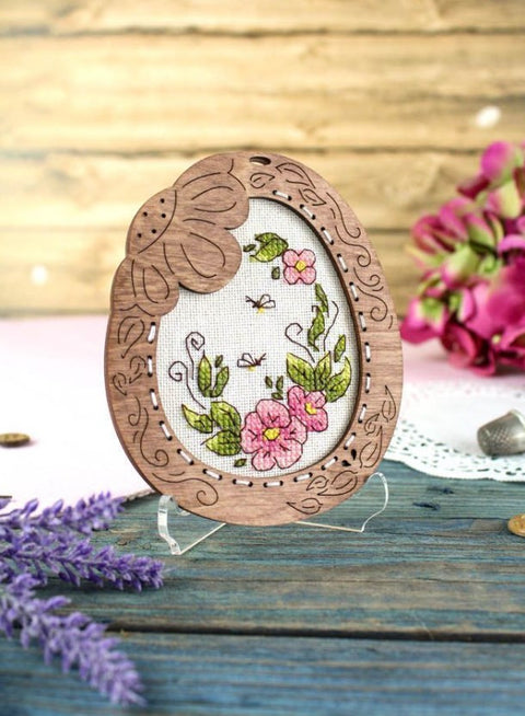 Medinis siuvinėjimo rėmelis ovalus su išraižytais ornamentais, rožinėmis gėlėmis siuvinyje ant skaidraus stovo