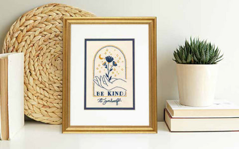 Įrėmintas plakatas su „Be Kind to yourself“ šviesiu kreminiu fonu ir tamsiai mėlyna grafika, elegantišku aukso rėmu