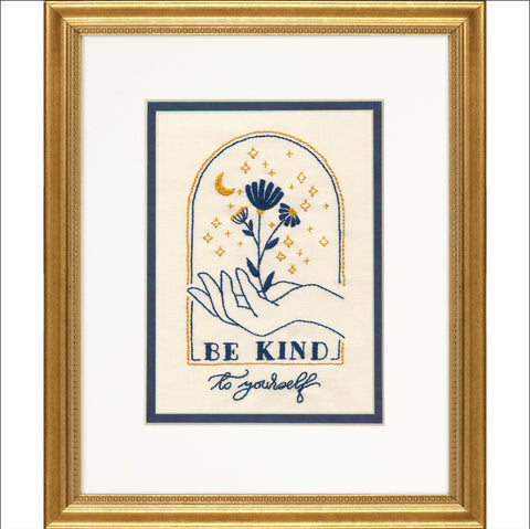 Rėminis siuvinys „Be Kind“ – mėlynos ir auksinės siūlės vaizduoja gėlę ir ranką ant smėlinio fono, su baltu kartonu ir auksiniu rėmu
