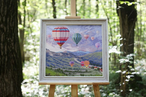 Paveikslas su karšto oro balionais baltame mediniame rėme, 40×40 cm, ant molberto miške — dekoratyvinis galerijos lauko variantas