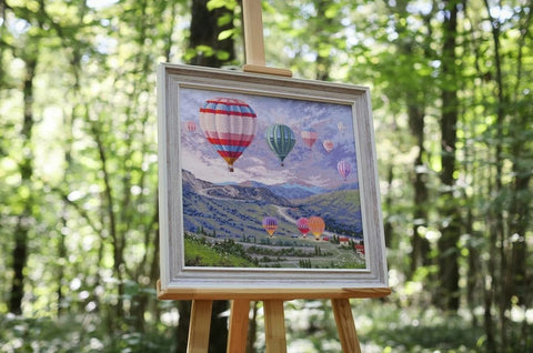 Paveikslas su spalvingais oro balionais, baltu mediniu rėmu 40×50 cm, pritvirtintas ant molberto miške, tinkamas svetainės akcentui