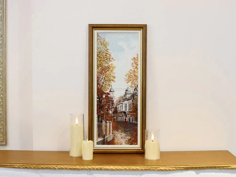 Siuvinėtas paveikslas rėmelyje, aukštas siauras rudeniškos gatvės motyvas su aukso rėmu, eksponuotas ant židinio tarp žvakių