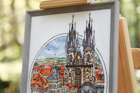 Siuvinėtas paveikslas su gotikinių katedros bokštų motyvu, pilko medinio rėmo ir stovo kompozicijos artimas planas