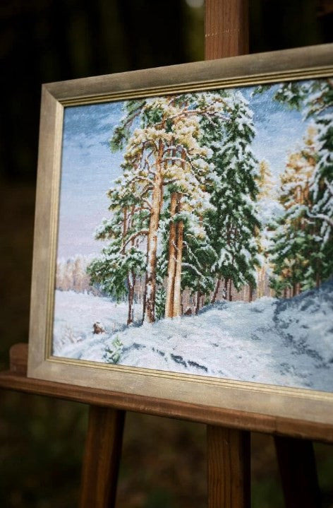 Siuvinėtas paveikslas: rėmuotas žieminis pušynas baltame sniege, aukso rėmas, nufotografuotas iš kampo ant medinio stovo