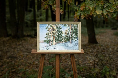 Paveikslas žiemiškas peizažas su auksiniu rėmu (40×60 cm), eksponuotas ant medinio stovo lauke miško fone