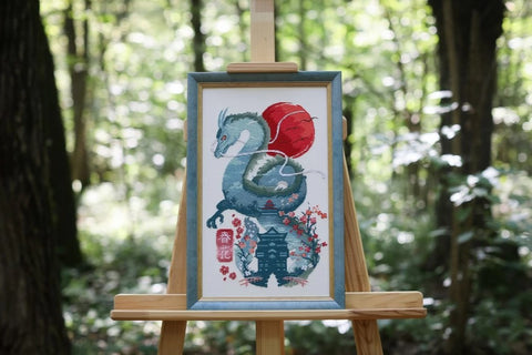 Siuvinėtas paveikslas su žaliu drakonu, raudonu saulės disku ir japonų paviljono motyvais, įrėmintas ant medinio stovo miške