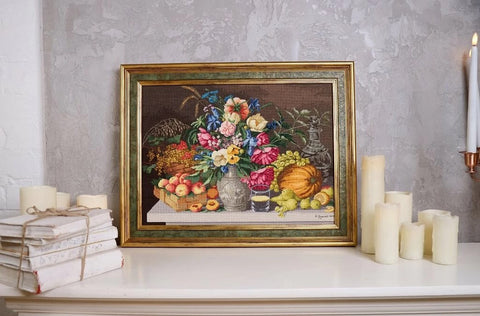 paveikslas rėmuotas gėlių ir vaisių reprodukcija auksiniame rėme, ~50×40 cm, ant baltos konsolės prieš pilką sieną šalia žvakių