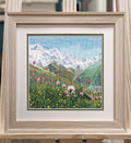 Siuvinėtas paveikslas šviesiame mediniame rėme 30×30 cm, alpių pieva su rausvomis gėlėmis ir snieguotais kalnais, ant molberto