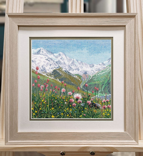 Siuvinėtas paveikslas šviesiame mediniame rėme 30×30 cm, alpių pieva su rausvomis gėlėmis ir snieguotais kalnais, ant molberto