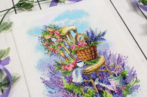 Siuvinėtas paveikslas kryželiu, medinė kėdė su gėlių krepšiu ir levandomis, pastelinės violetinės spalvos, kampinis artinys