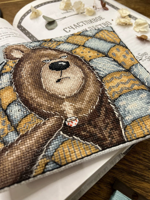 Siuvinėtas užrašų knygos viršelis su rudu lokio motyvu, mėlyno‑geltono patchwork raštu ir ryškiomis rankų siūlėmis