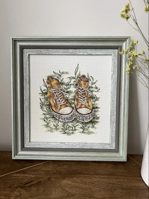 Siuvinėtas paveikslas geltonu kedų motyvu su pilku medžio rėmu (apie 30×30 cm), centre ant medinio stalo