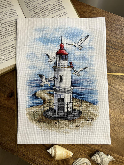 Siuvinėtas švyturys ant balto audinio su raudonu kupolu ir žuvėdromis, rankų darbo 21×30 cm jūrinė dekoracija ant medinio stalo, matoma su kriauklėmis ir atviru knygos kampu