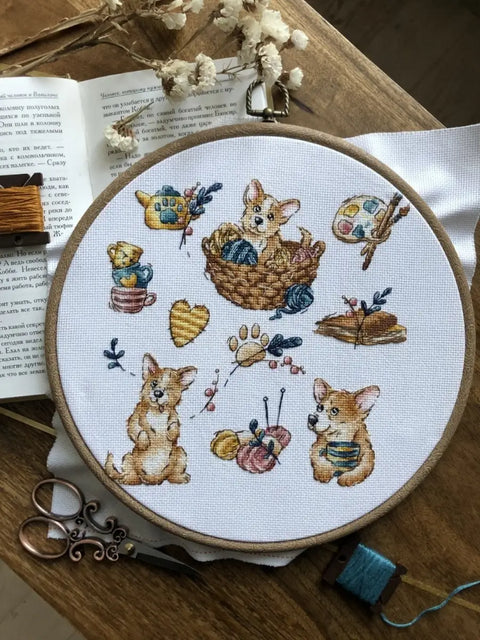 Kryželių siuvinėjimo rinkinys su corgi motyvais, mediniu ratu, siūlų kamuoliukais ir ant stalo esančiomis žirklėmis