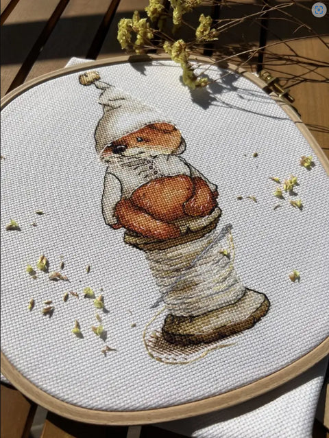 Siuvinys – rankų darbo meškiuko ornamentas su oranžinėmis detalėmis ant balto audinio, arti parodytas siūlų ritinys ir lankelis