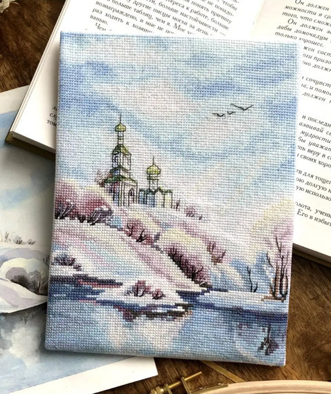 Siuvinėtas paveikslas žieminės bažnyčios motyvas, pasteliniai mėlyni ir rožiniai tonai, auksinės kupolų detalės, matoma rankų darbo tekstūra ant stalo