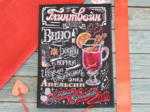 Siuvinėtas plakatas juodame audinyje su spalvotu glintveino receptu, stikliniu puodeliu ir cinamono lazdelėmis, parodytas iš viršaus su oranžiniu kaspinu ir raudona staltiese
