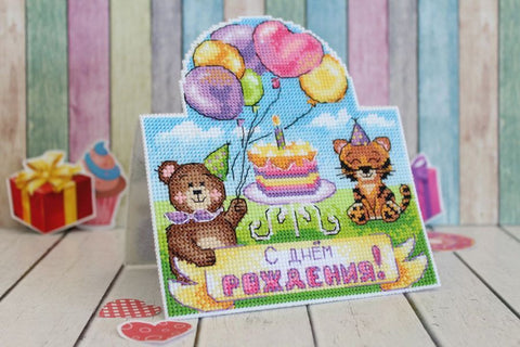 Siuvinėta atvirutė su gimtadienio meškiuku ir kate, spalvoti balionai bei tortas, stovinti ant stalo