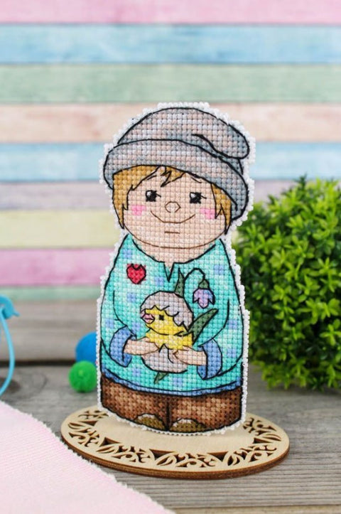 Siuvinėta figūrėlė, priekinė pusė: berniukas su viščiuku, pasteliniai atspalviai ir medinė atrama.