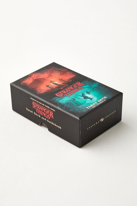 Stranger Things tarot kortų rinkinys juodoje dėžutėje su raudonais ir žalsvais viršelio piešiniais ir pridedamu vadovu, nukreiptas kampiniu rakursu