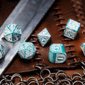 RPG kauliukai tirkiai‑sidabriniai, graviruotų 7 dalių rinkinys, artimas kadras ant odinio žaidimo kilimėlio su metaline grandinėle